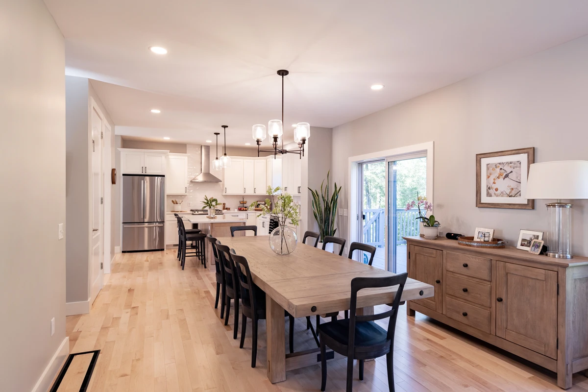 A long dining table running toward an open white kitchen, on light maple floors