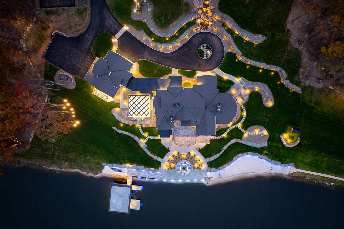 An aerial view at dusk of a lakefront home’s lit paths and gardens