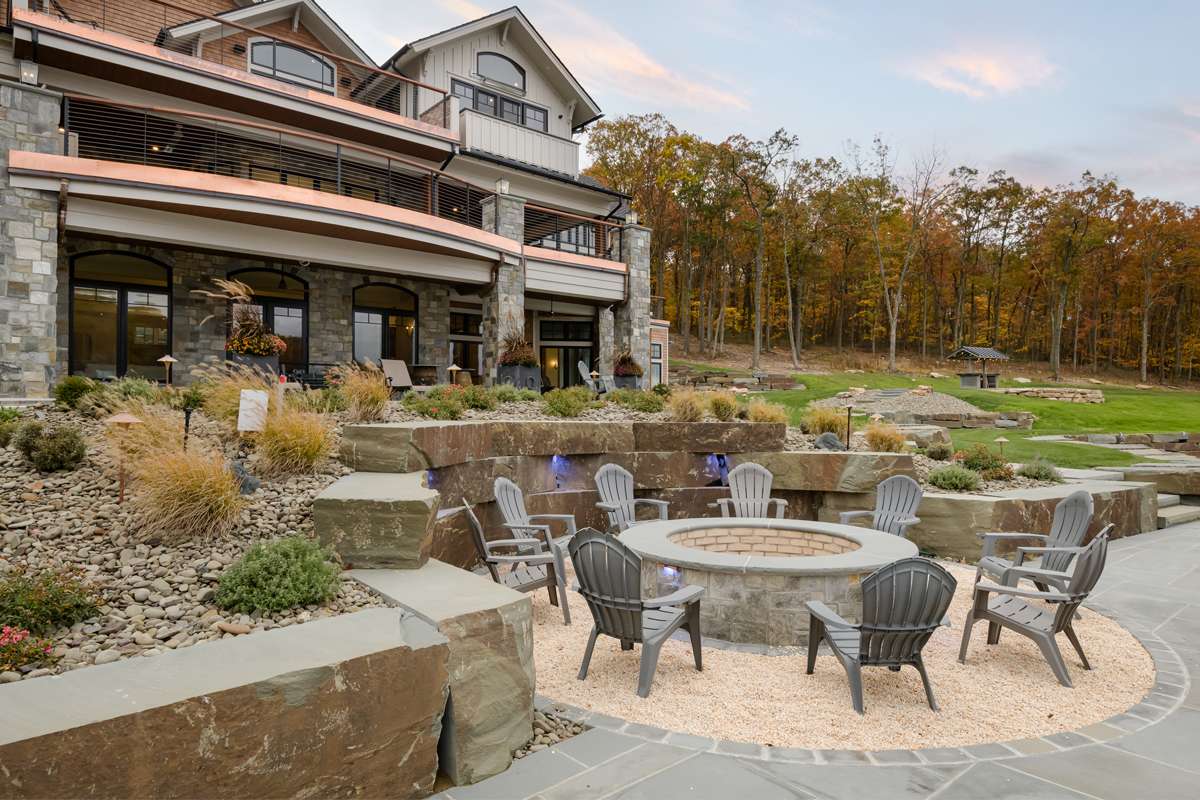 A stone fire pit terrace and planted walls behind a new lake home