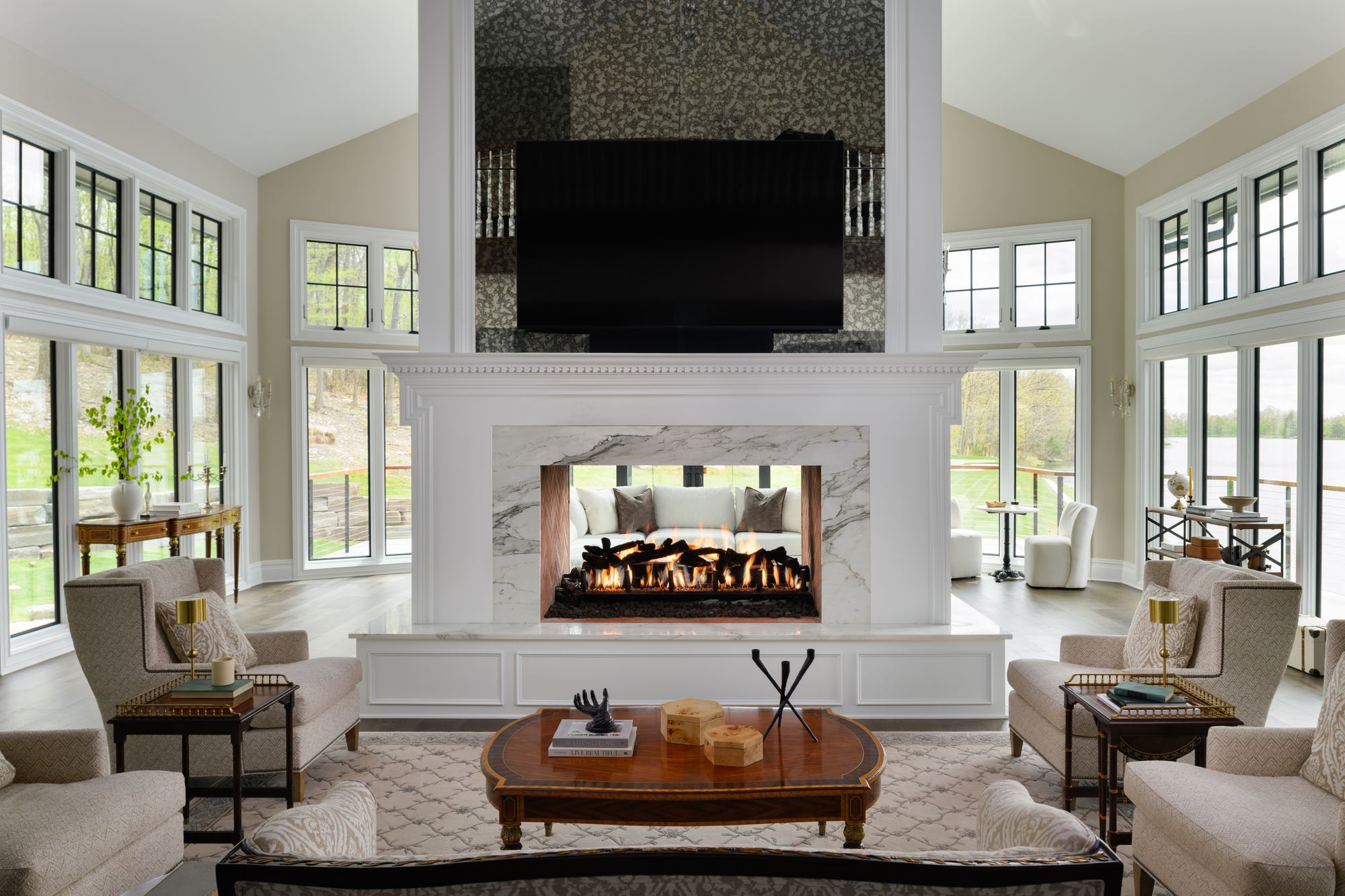 A great room with a marble double-sided fireplace under a vaulted ceiling, walls of windows on both sides