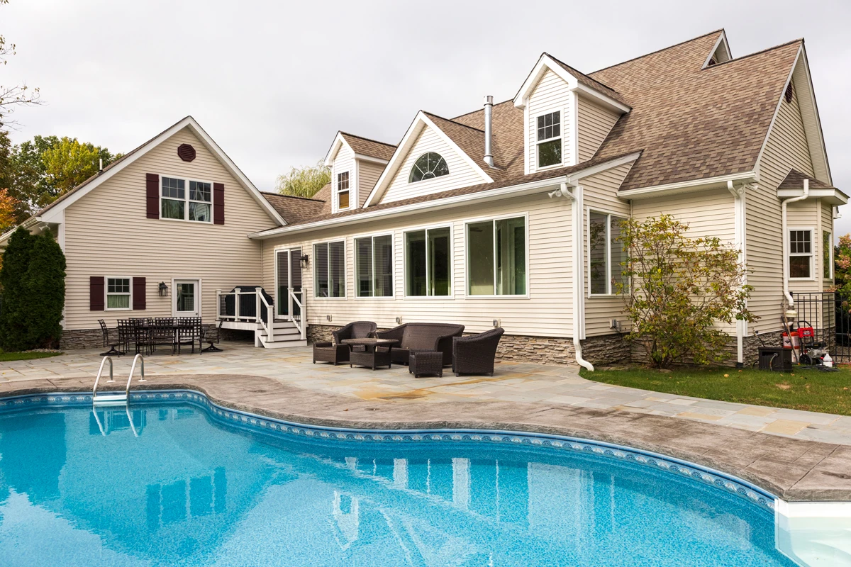 A sunroom addition across the back of a Cape Cod home, above a bluestone patio and an in-ground pool