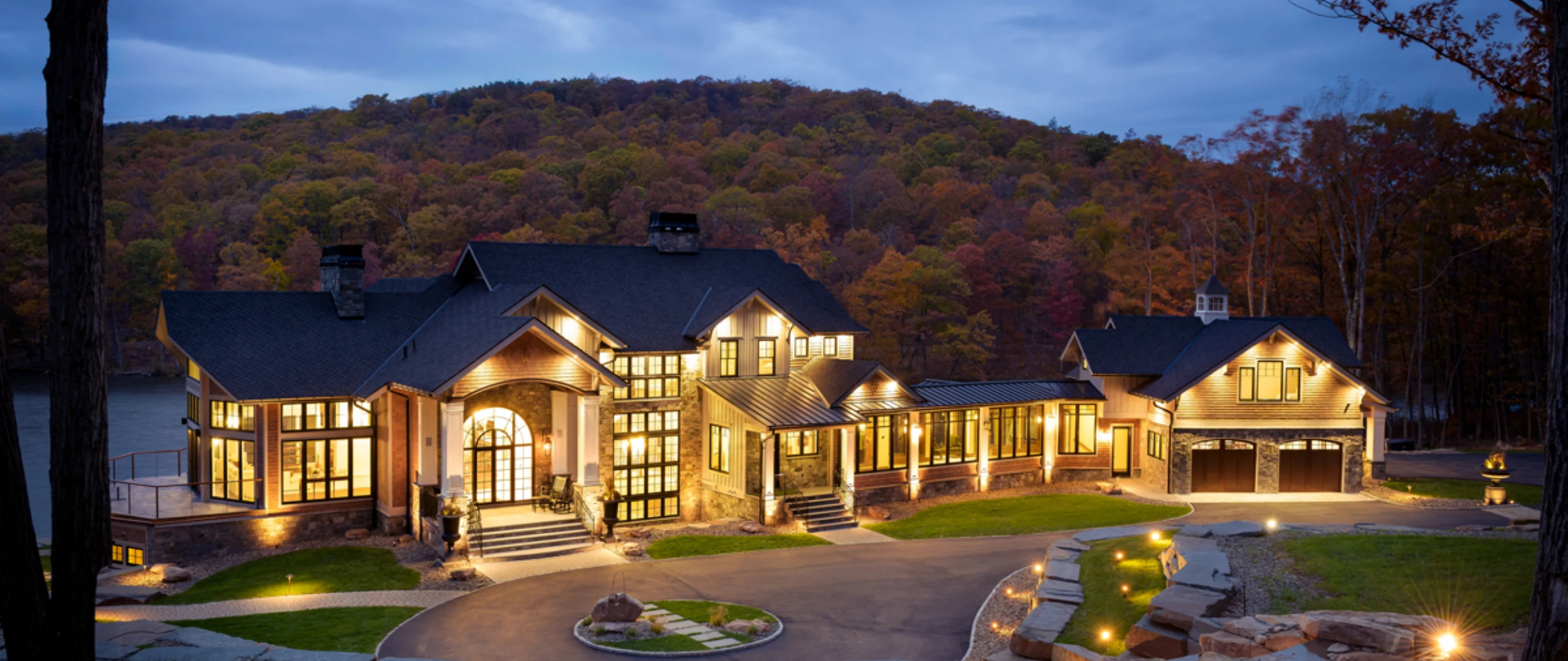 A lakefront custom home lit at dusk, stone walls and lawn stepping down to the drive
