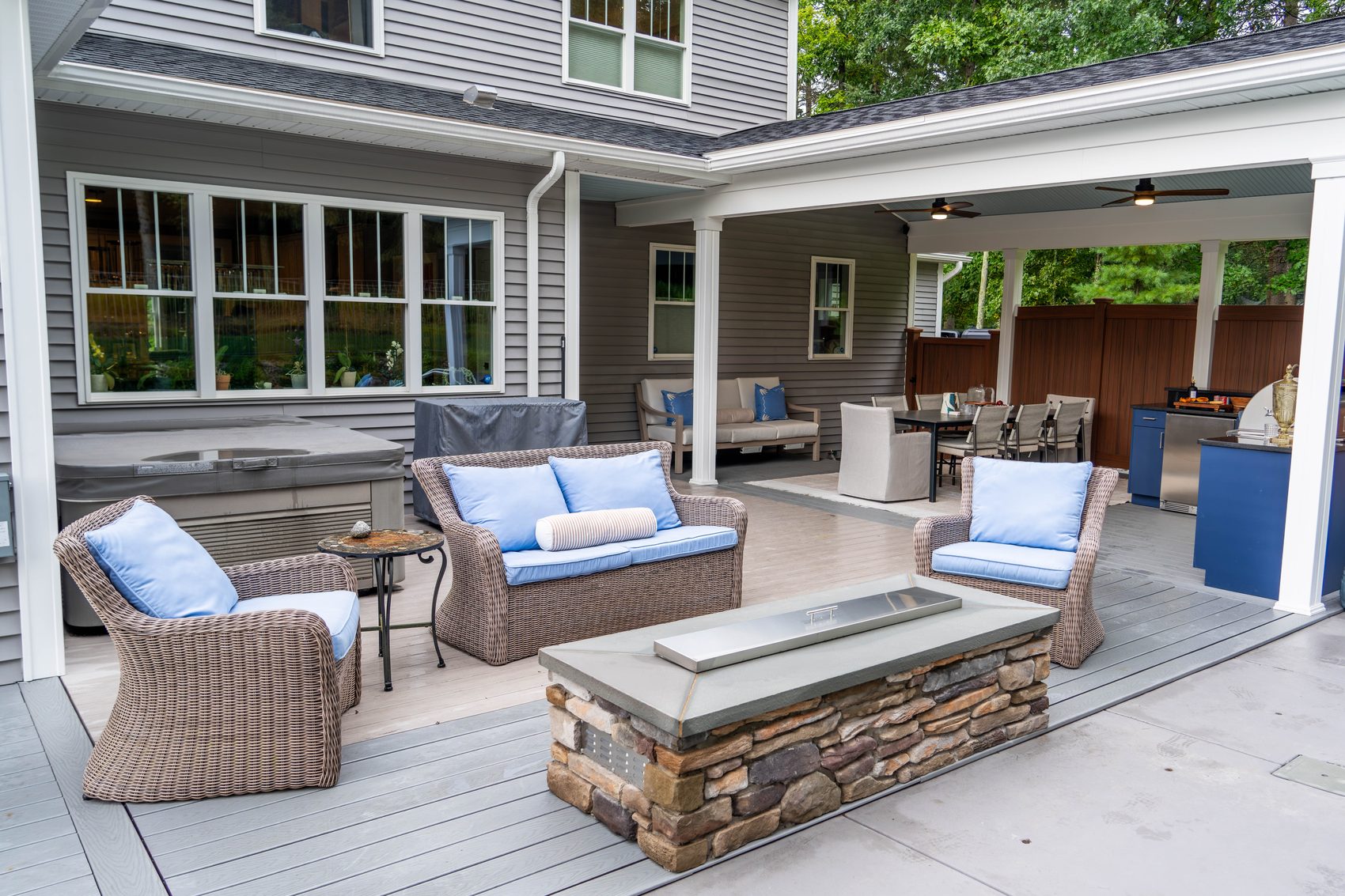 A covered composite deck with wicker seating around a stone fire table, the outdoor kitchen behind