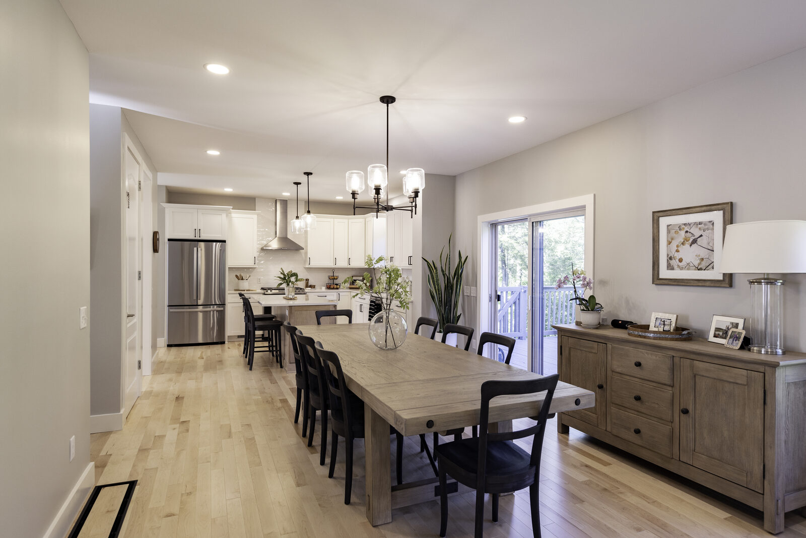 A long dining room with a farmhouse table, black chairs and a chandelier, open to a white kitchen