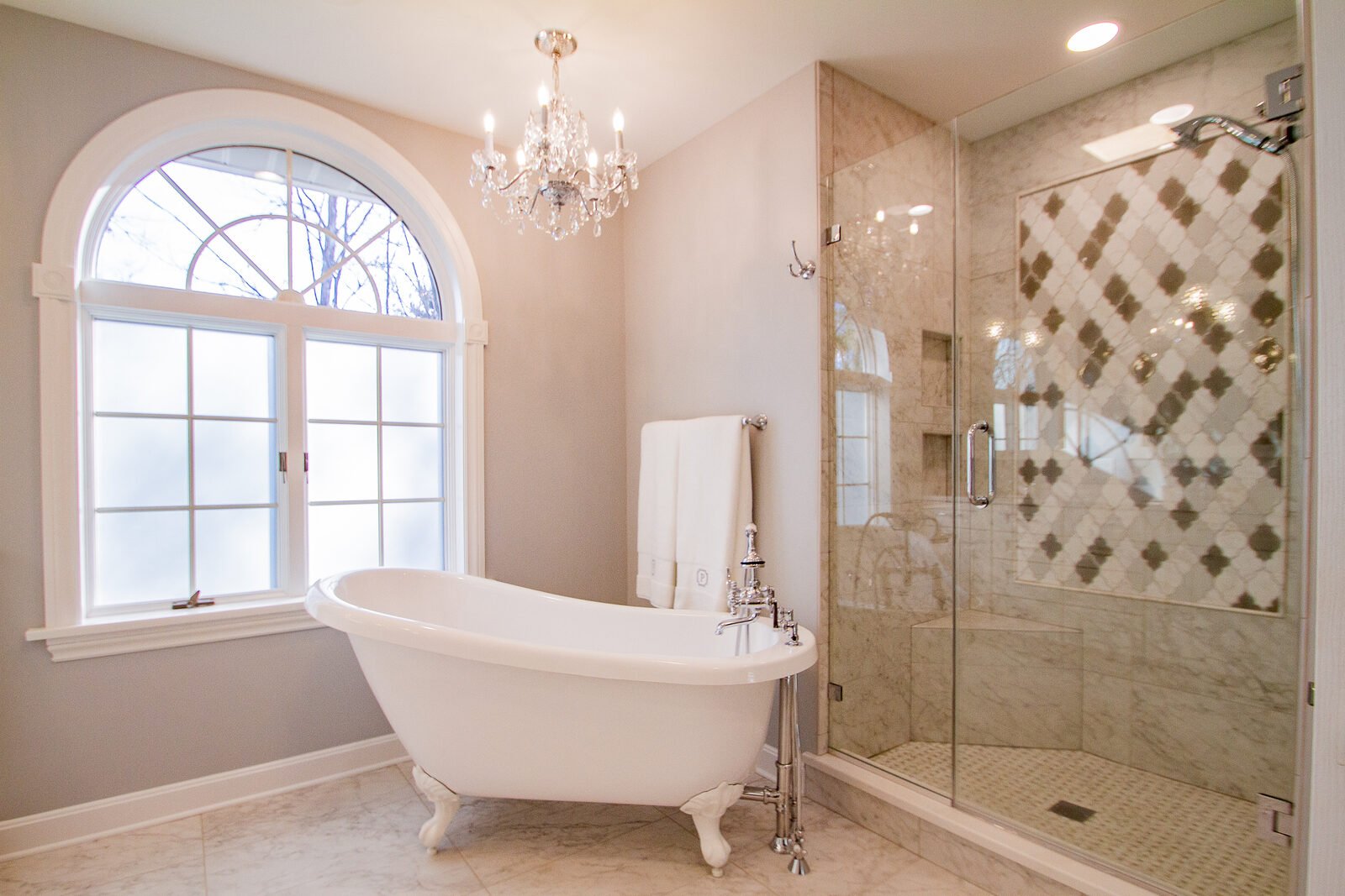 A primary bathroom with an arched window, a clawfoot tub, a crystal chandelier and a glass shower with patterned tile
