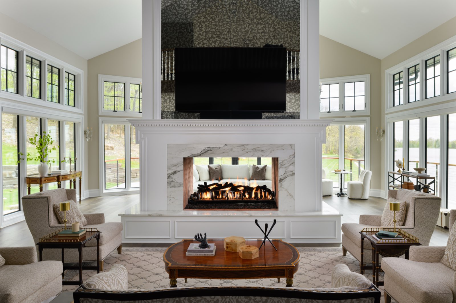 A vaulted great room with a see-through marble fireplace, walls of black-framed windows and armchairs around a wood coffee table