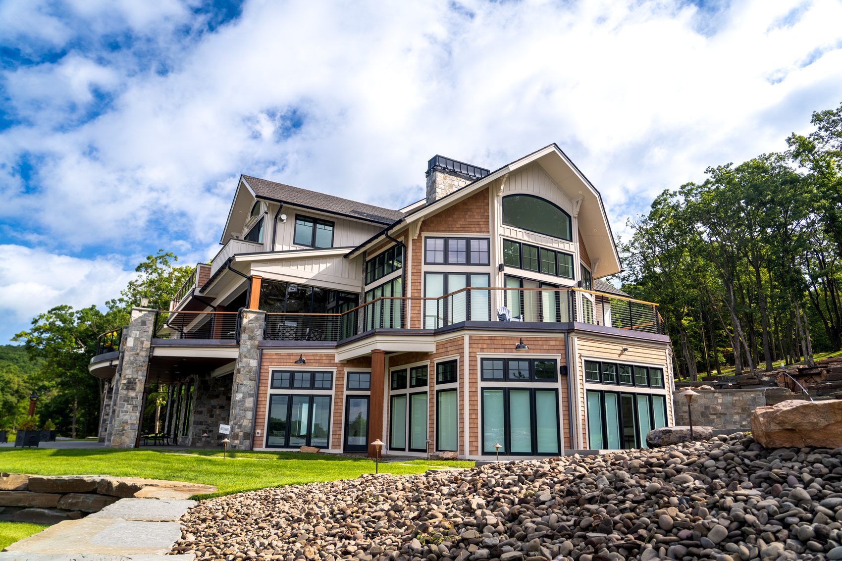 A shingle-and-stone lake house with tall arched windows and a wraparound deck above a rocky slope